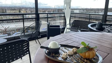 Una taula de menjador amb un plat de postre, un bol de gelat i una calabossa decorativa en un balcó amb vista a un paisatge urbà.