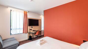 Ein Hotelzimmer mit Bett, Schreibtisch mit Stuhl, Wandfernseher und einem Fenster mit Vorhängen.