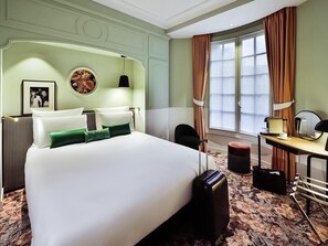 Hotelkamer met een groot bed, een bureau met stoel, een klein tafeltje, een spiegel en een raam met gordijnen.
