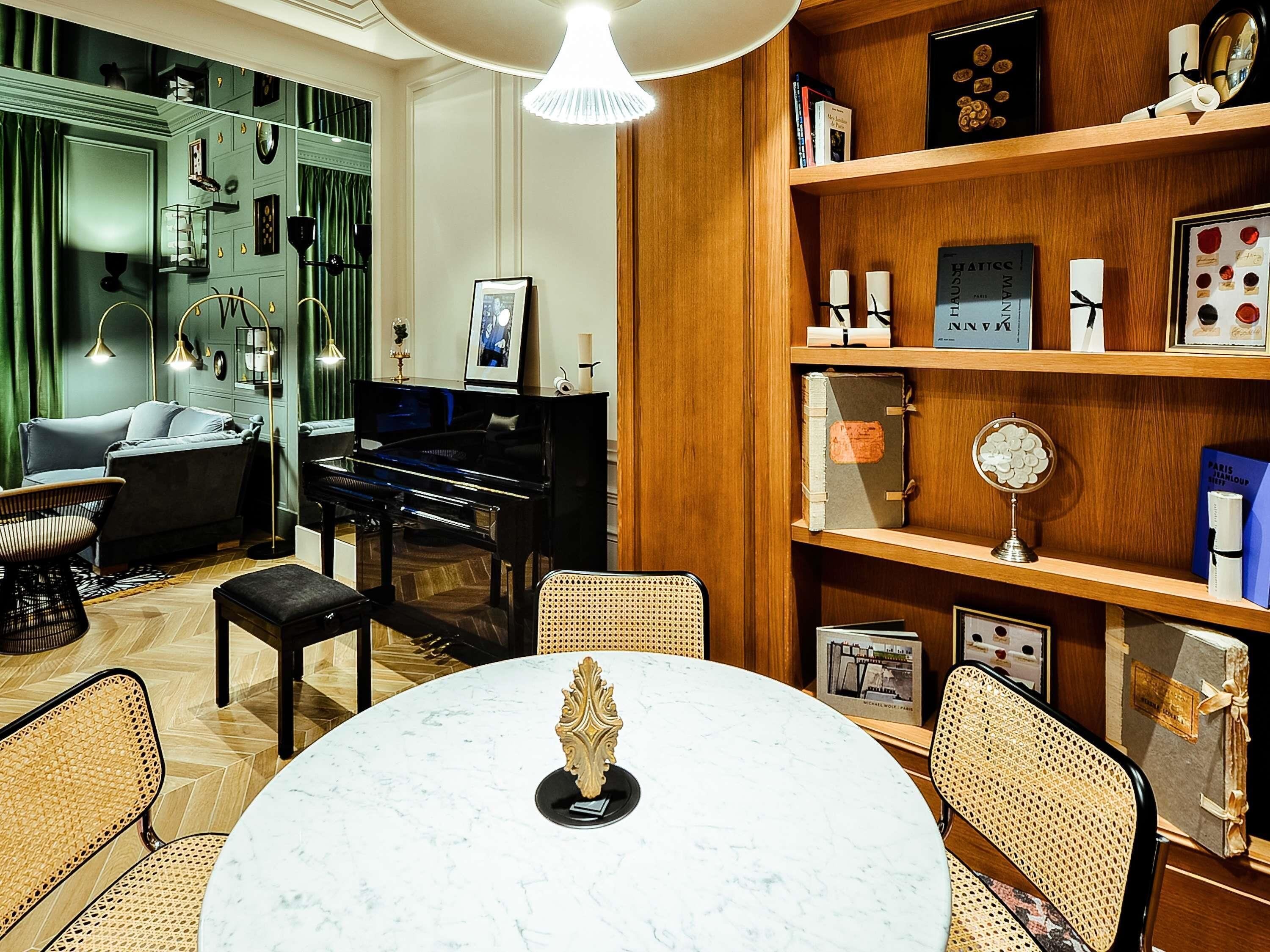 A room with a piano, a round table with a decorative centerpiece, wicker chairs, a bookshelf with various items, and a green wall with a mirror.