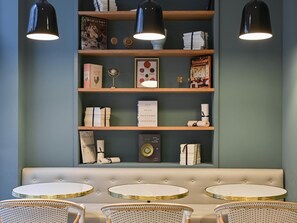 Een zithoek met een gestoffeerde bank en ronde tafels, een boekenkast met boeken en decoratieve objecten, en hanglampen.