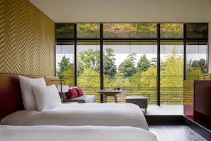Quarto de hotel moderno com uma cama grande, uma mesinha e vista para árvores através de amplas janelas.