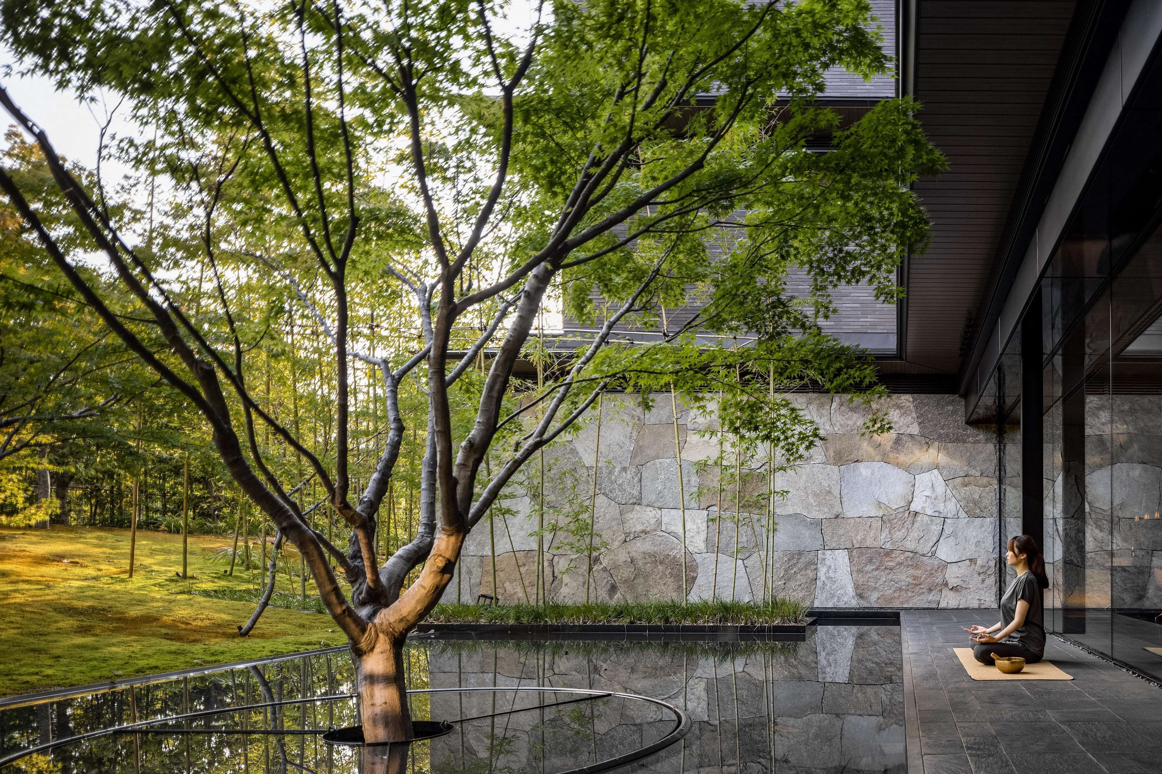 A person meditating on a mat in a modern outdoor setting with a stone wall and a tree.