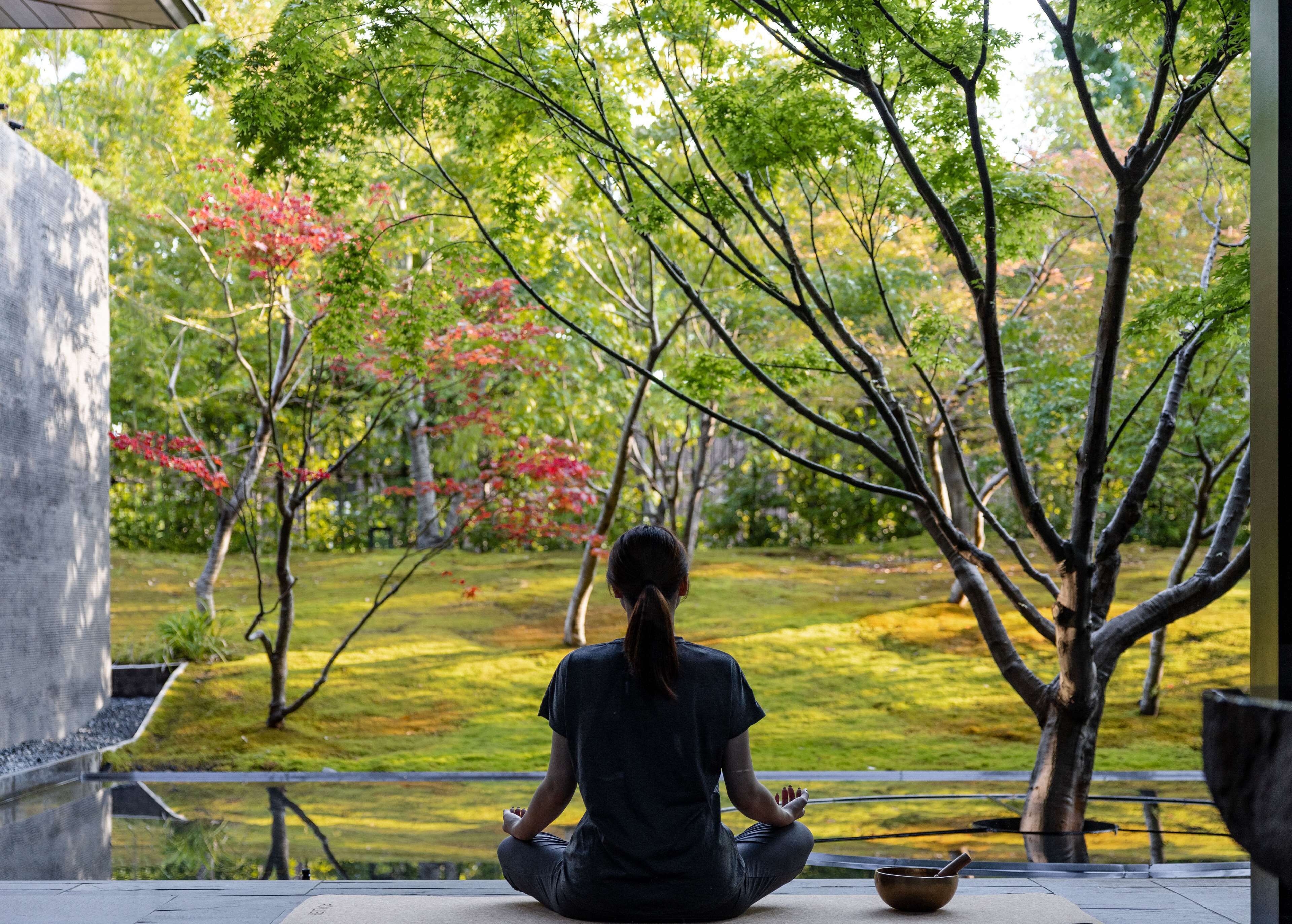 Una persona meditant en un entorn exterior tranquil, amb arbres i un estany.