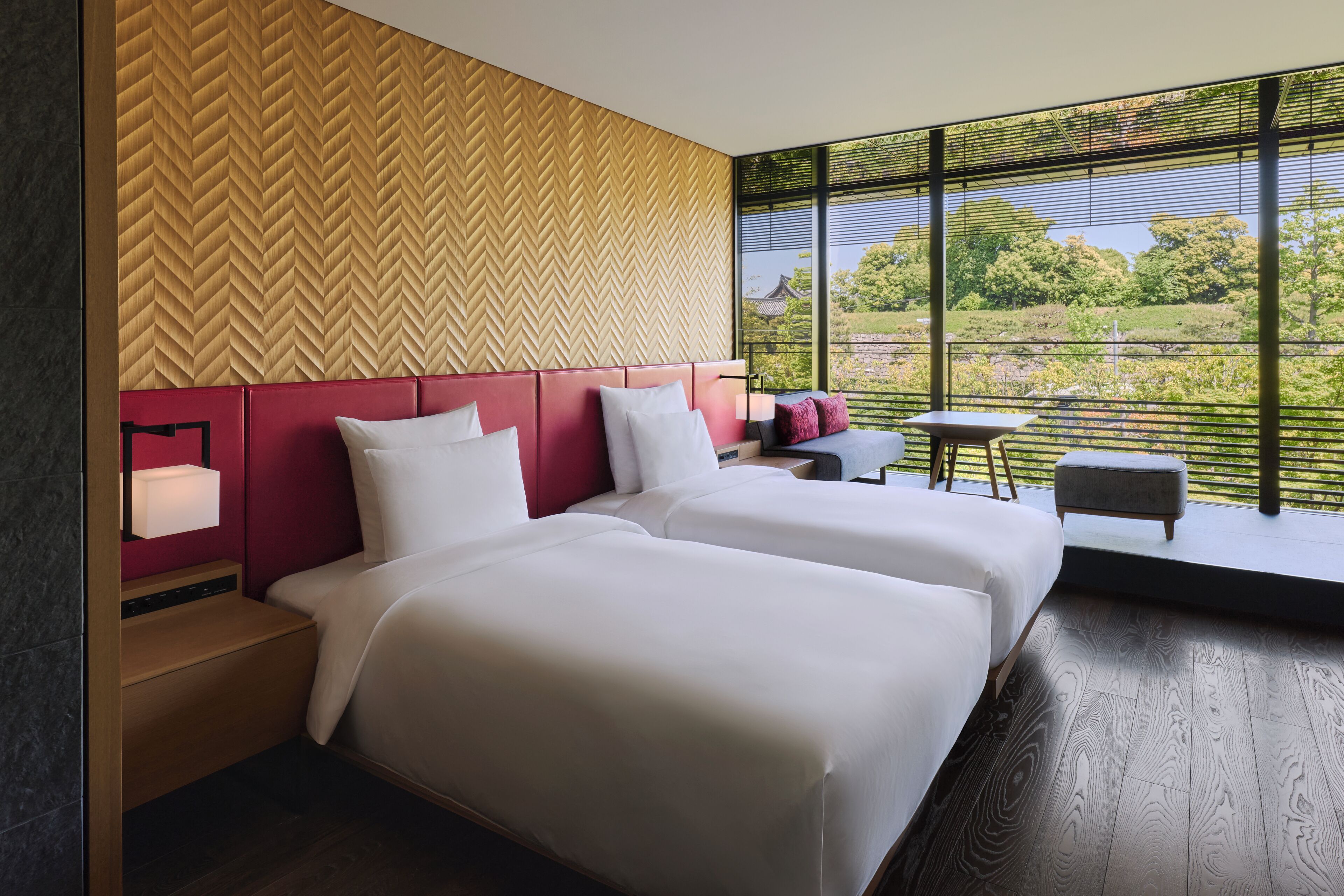 A modern hotel room with two beds, a red headboard, a large window with a view of greenery, and a small table with a chair.