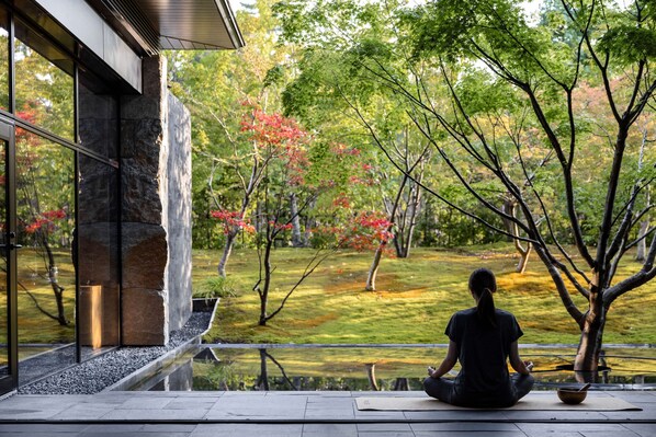 Una persona medita in un ambiente naturale tranquillo, con vista su un giardino e un edificio.