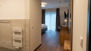 Una habitació d'hotel moderna amb sòl de fusta, un llit amb capçalera blanca, una tauleta de nit amb una làmpada i un mirall gran a la paret.