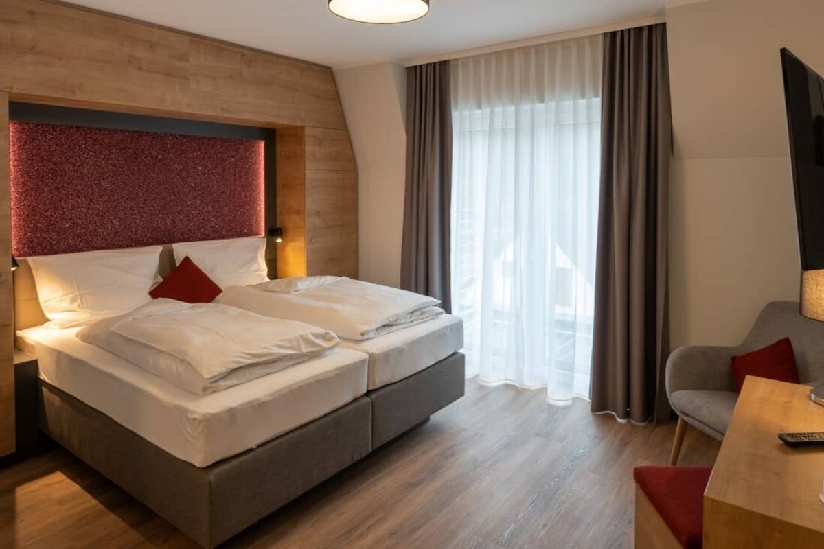 Una habitación de hotel moderna con una cama grande, cabecera de madera, un sofá gris y una televisión.