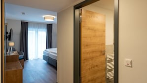 Habitació d'hotel amb una porta de fusta, un llit, un escriptori amb una làmpada i vista a l'exterior a través d'una finestra amb cortines.
