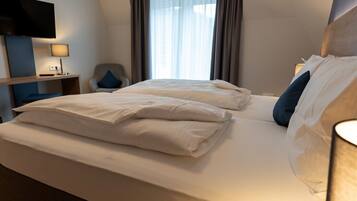 A hotel room with a large bed, a TV mounted on the wall, a desk with a lamp, and a window with curtains.