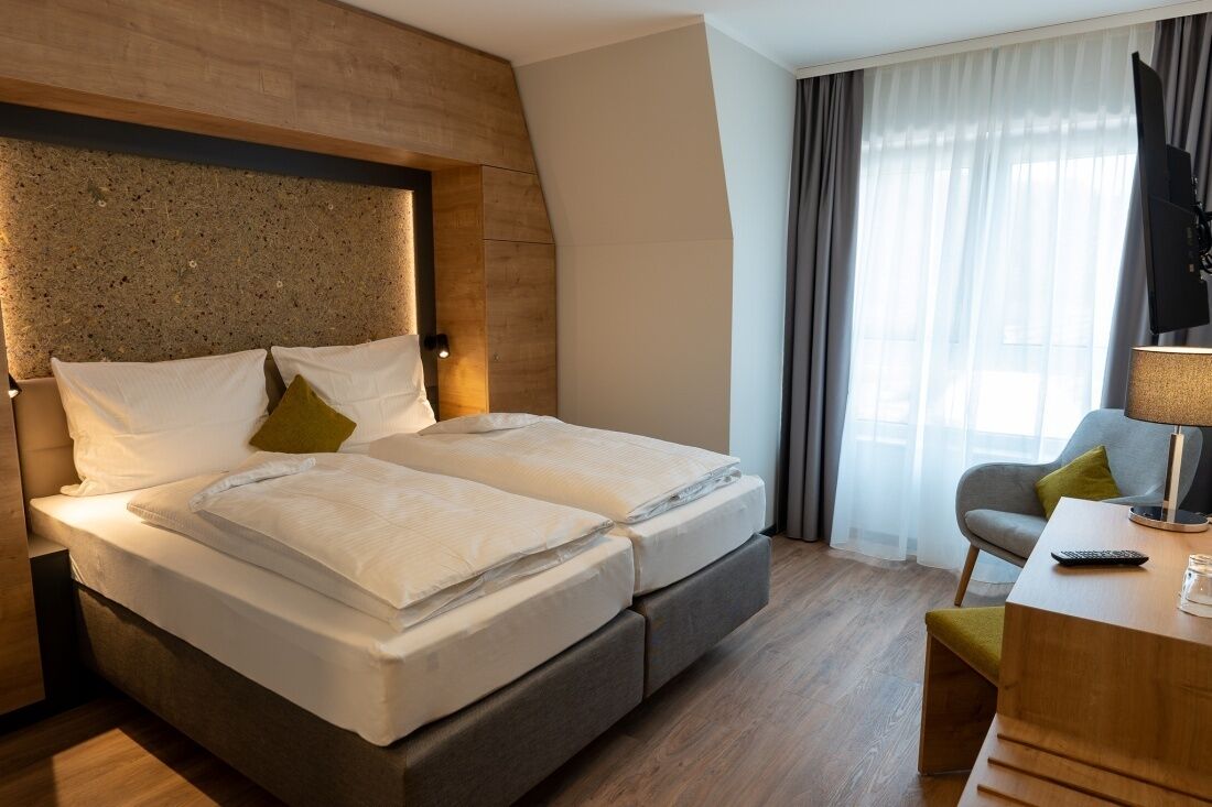 Una cama doble con cabecera de madera, una mesita de noche con lámpara, y una silla con cojín.
