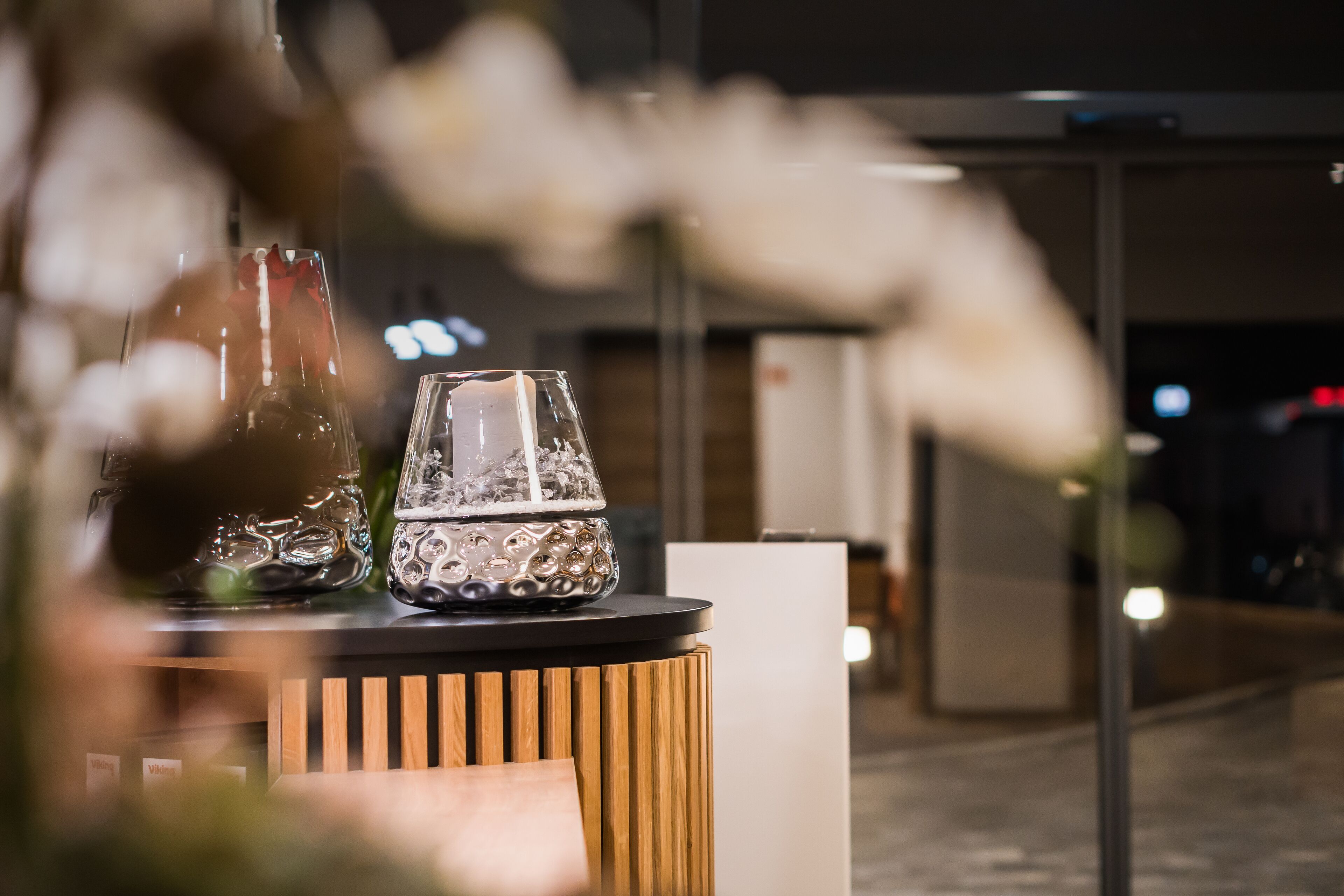 A table with two glass vases and a candle, set against a modern interior with a wooden accent and a glass partition in the background.