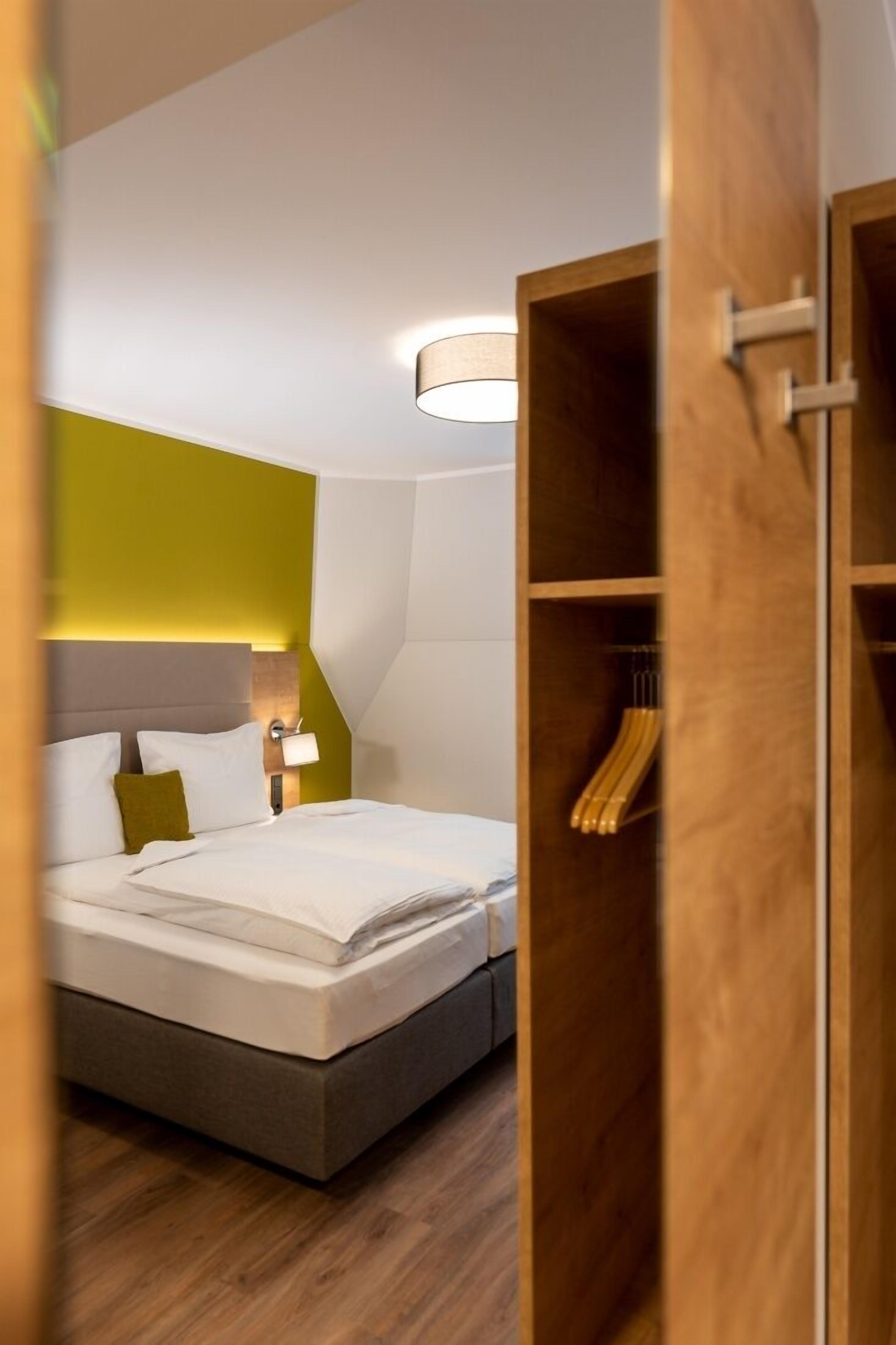 A modern hotel room with a neatly made bed, a wooden wardrobe, and a wall-mounted light fixture.
