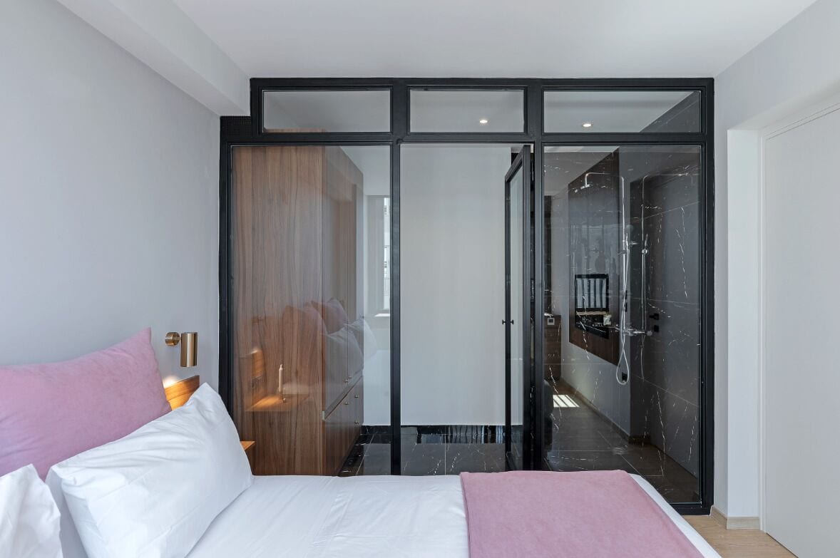 Une chambre d’hôtel moderne avec un lit, un plaid rose, une armoire en bois et une porte en verre donnant sur une salle de bain avec douche.