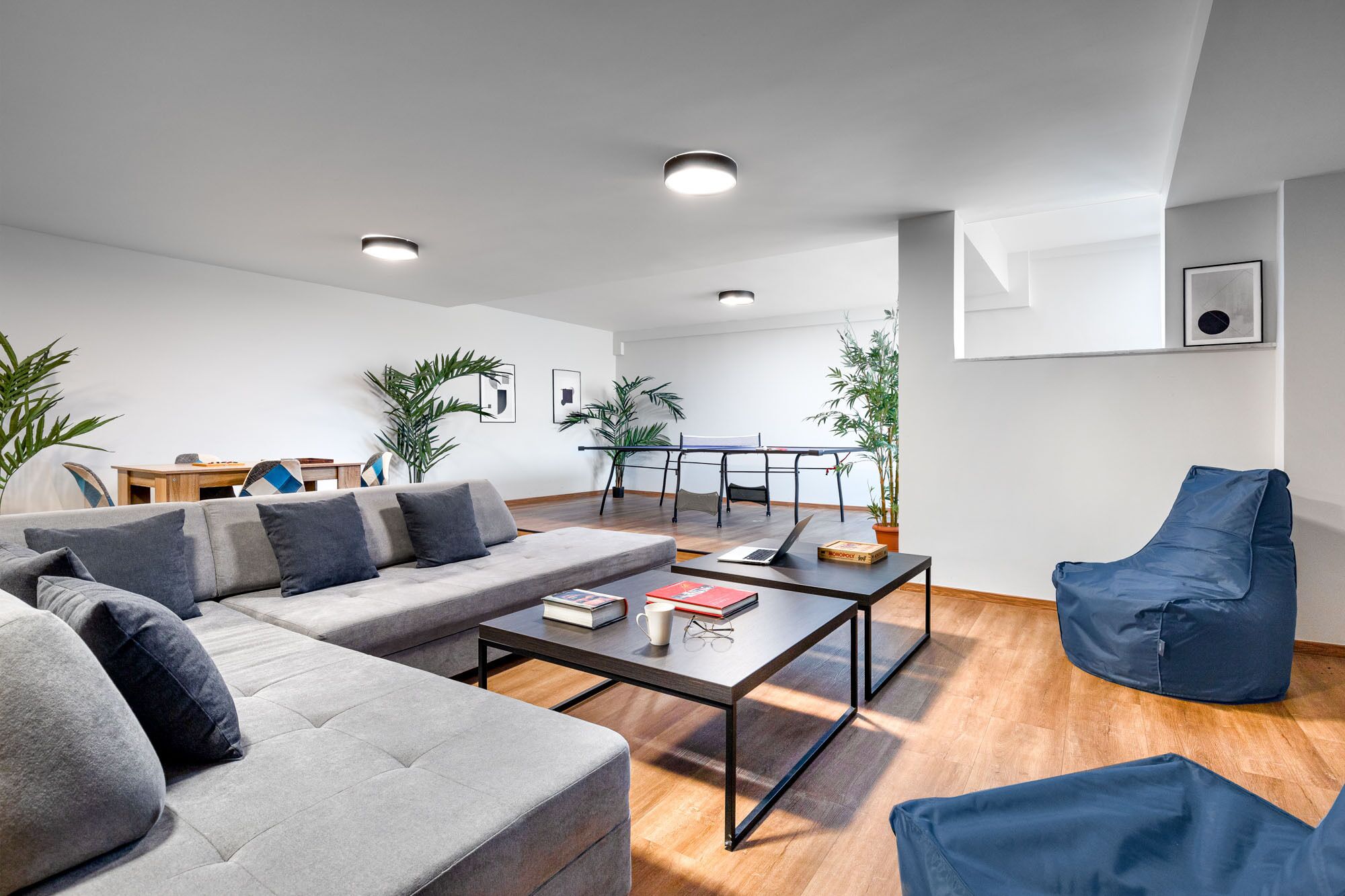 Una sala de estar moderna con un sofá gris, una mesa de centro y una silla azul. Se ve una mesa de ping pong al fondo y varias plantas en macetas.