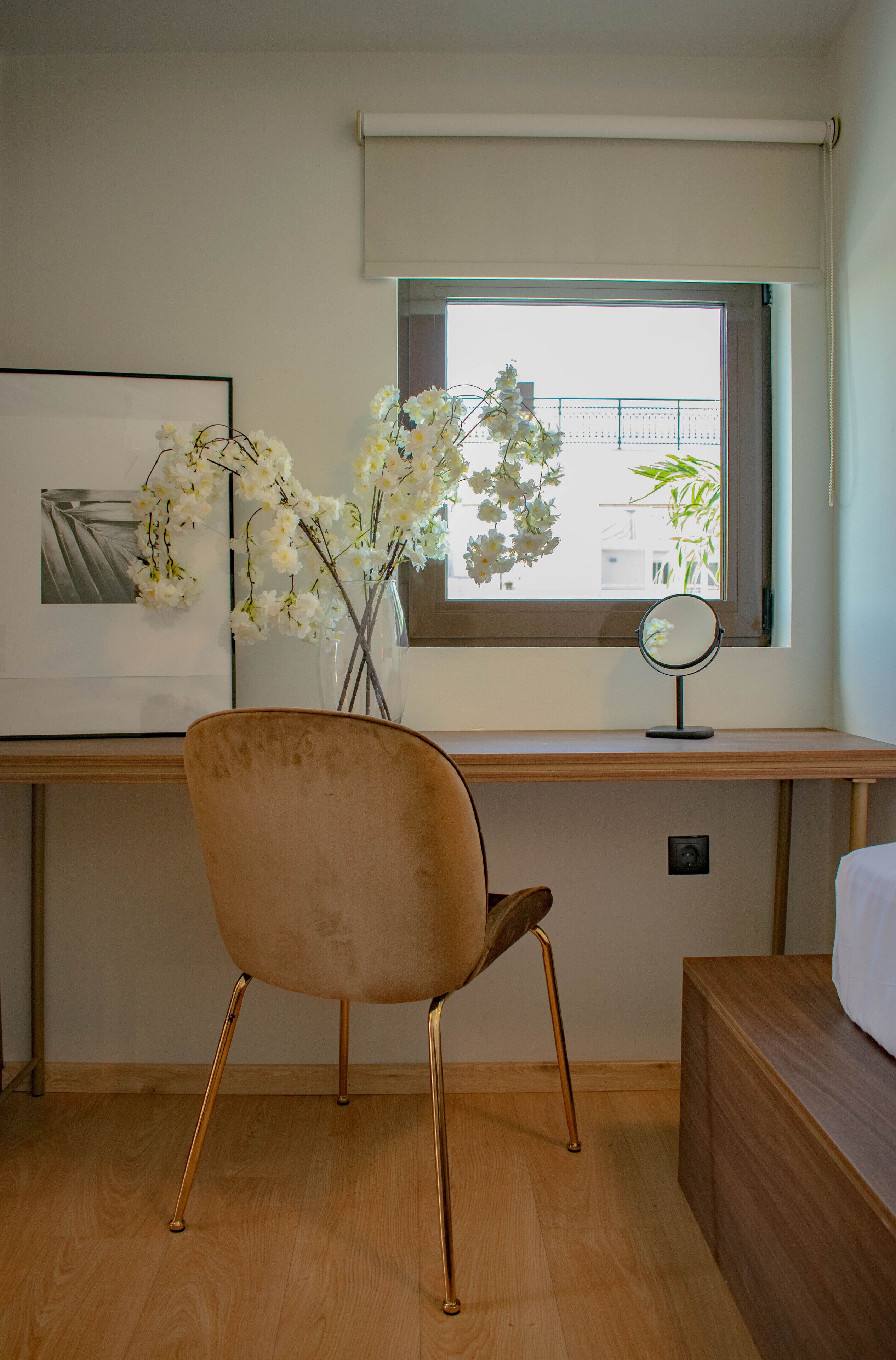 A bedroom with a wooden desk, a chair, a bedside table, a bed, a window with a view of buildings, and a vase with flowers.
