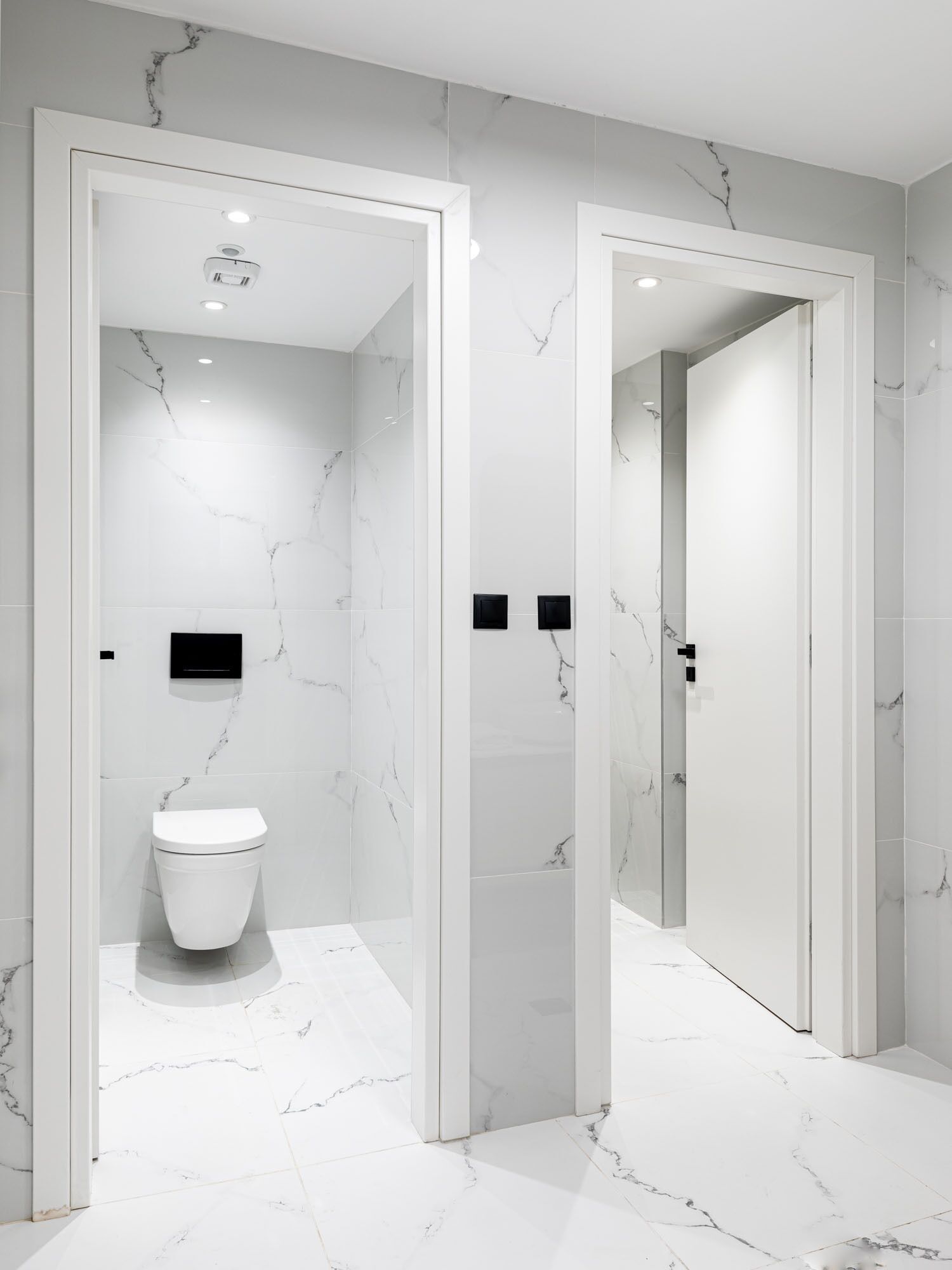 Een moderne badkamer met witte marmeren wanden en vloeren, een toilet en twee zwarte, vierkante objecten aan de muur.