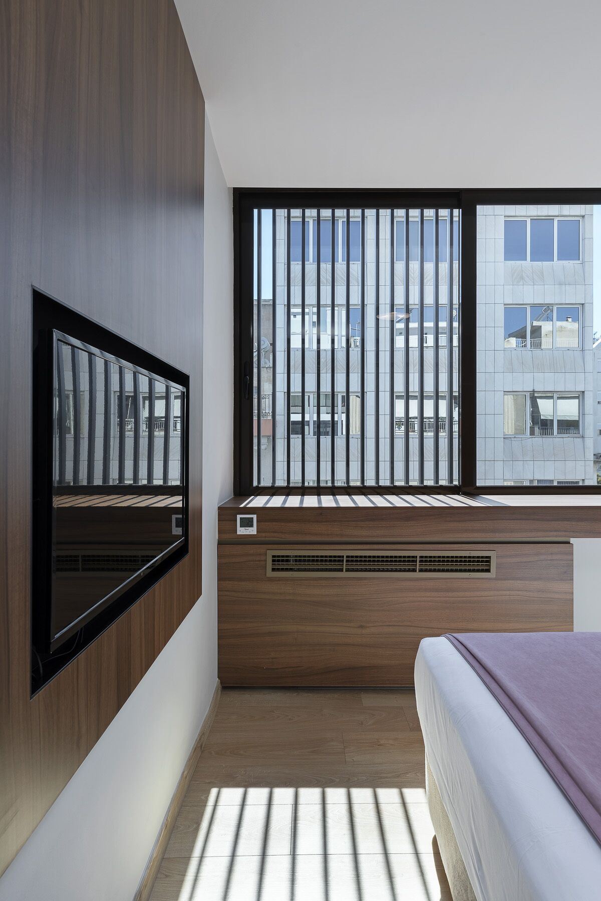 A modern bedroom with a large window, built-in TV, and a bed with a purple blanket.