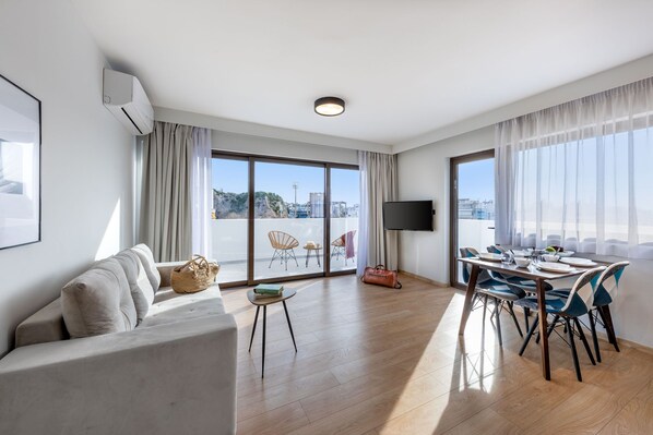 A modern living room with a sofa, a dining table with chairs, a TV, and large windows with a city view.