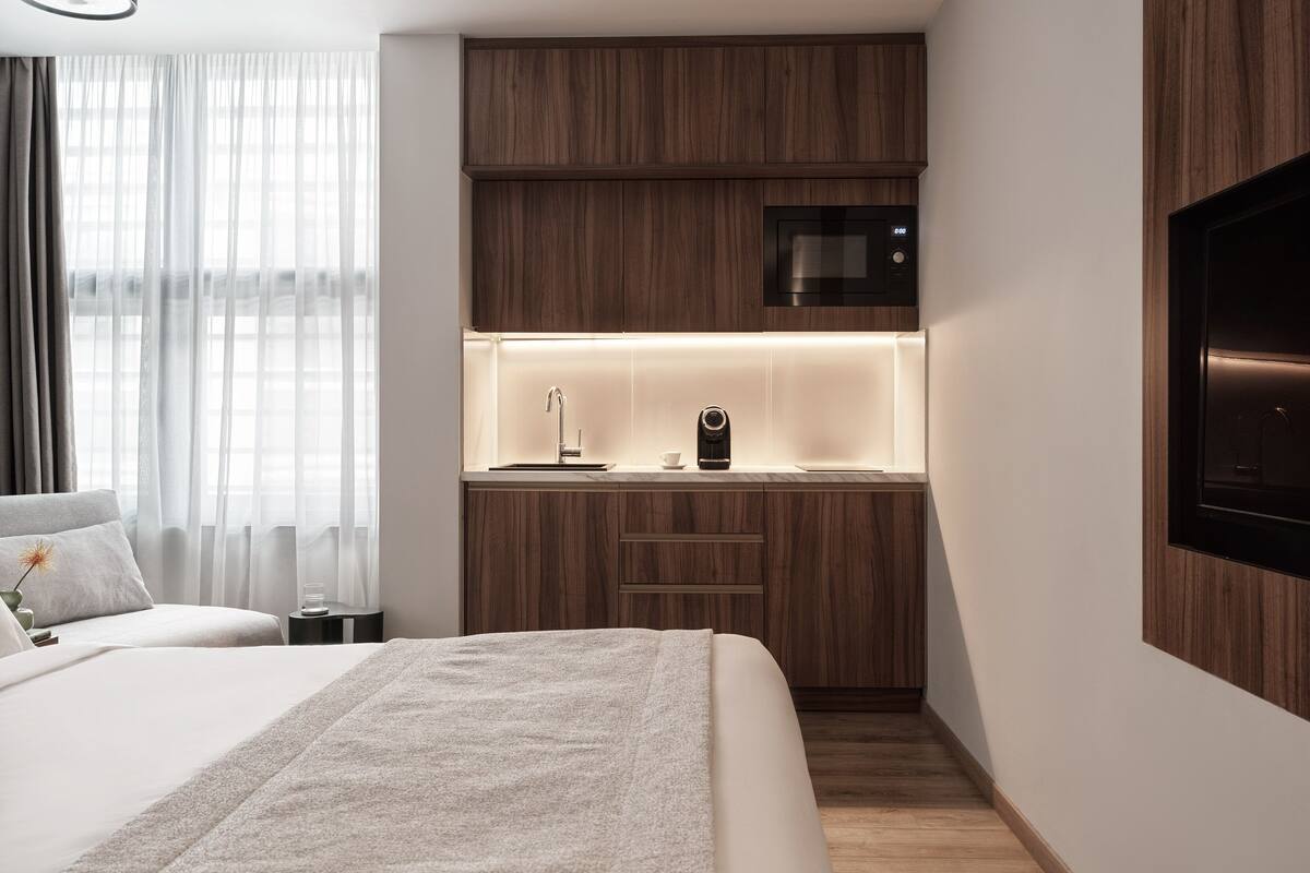 A modern hotel room with a bed, a bedside table, a nightstand, a built-in kitchen with a microwave, and a TV mounted on the wall.