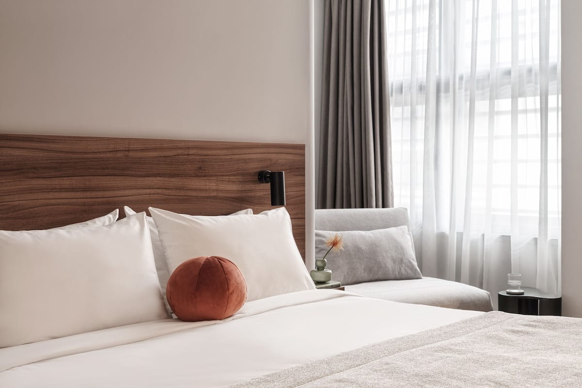 A modern hotel room with a wooden headboard, a large bed with white and beige bedding, a red accent pillow, and a side table with a vase.