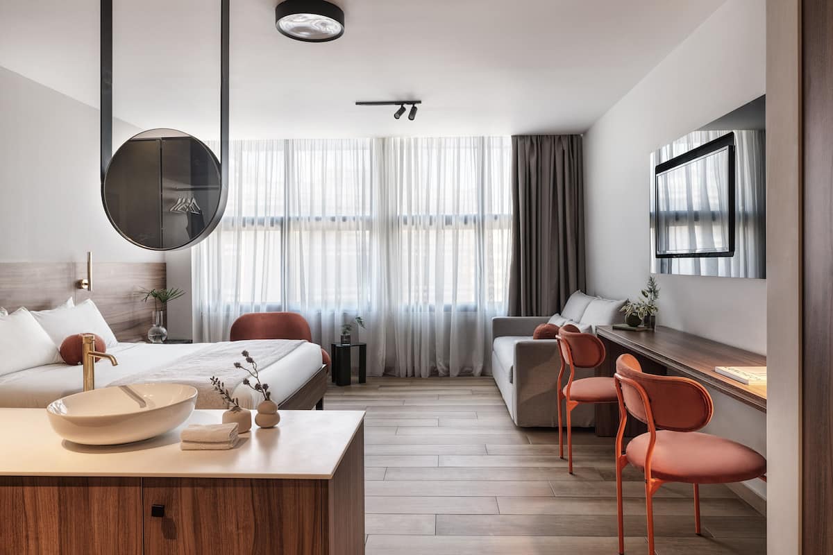 A modern hotel room with a bed, a desk, a chair, a sofa, and a TV mounted on the wall.