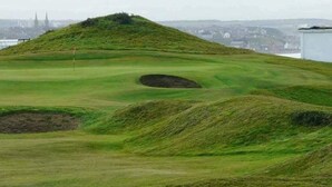 Ein Golfplatz mit Bunker, Fahne und einer in der Ferne liegenden Stadtansicht.