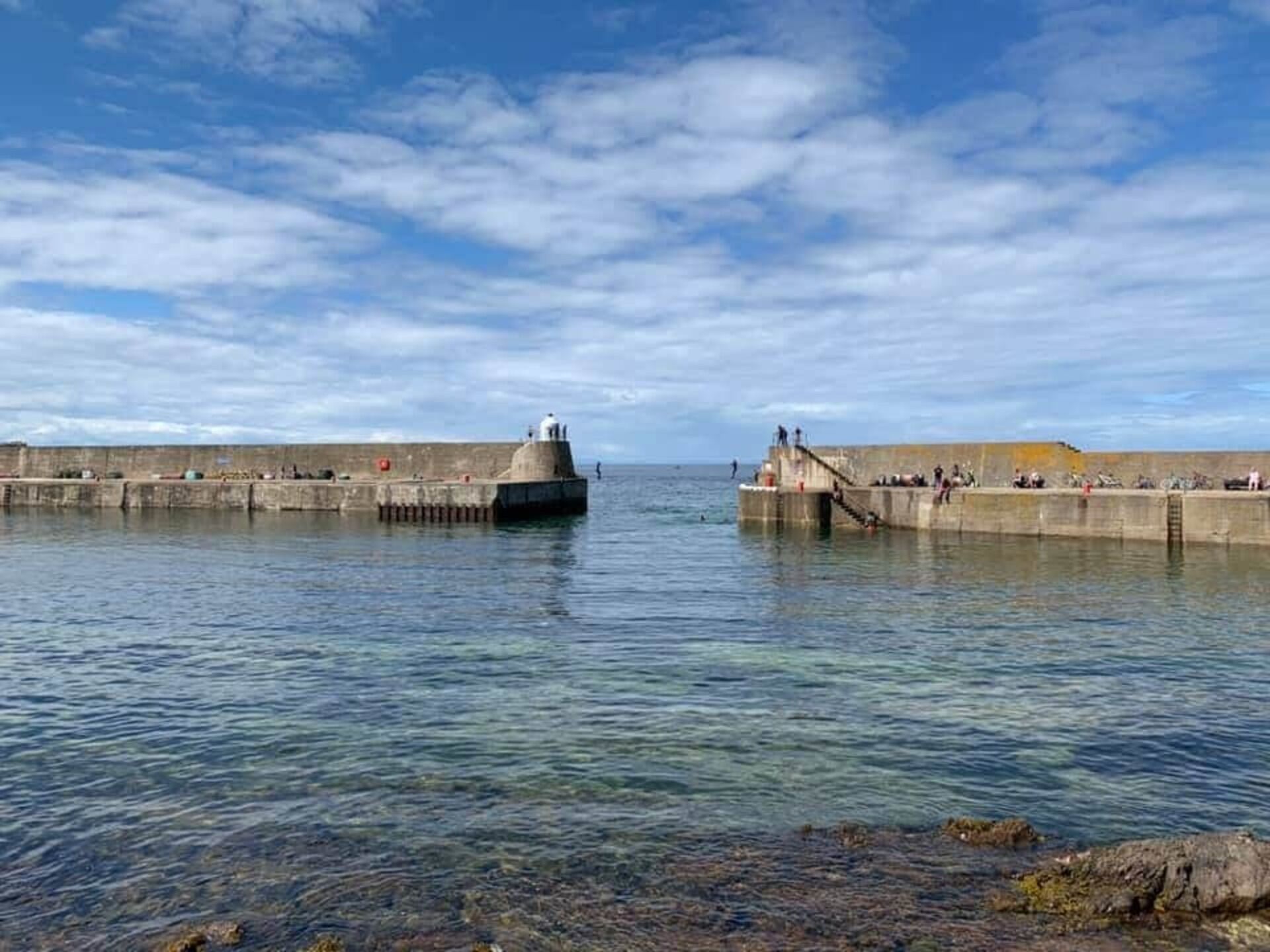 A coastal pier with a lighthouse, clear water, and a stone wall.