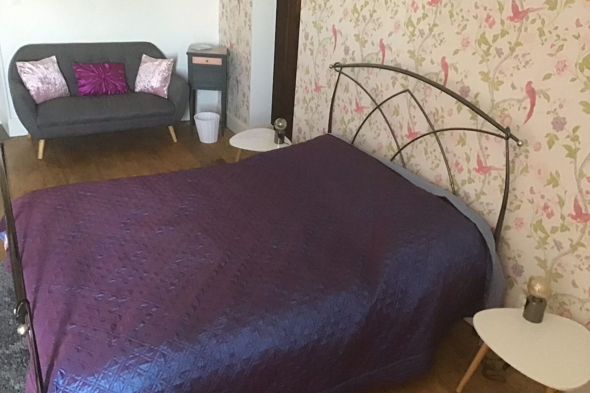 A bedroom with a bed, a nightstand, a small table, a sofa, and floral wallpaper.