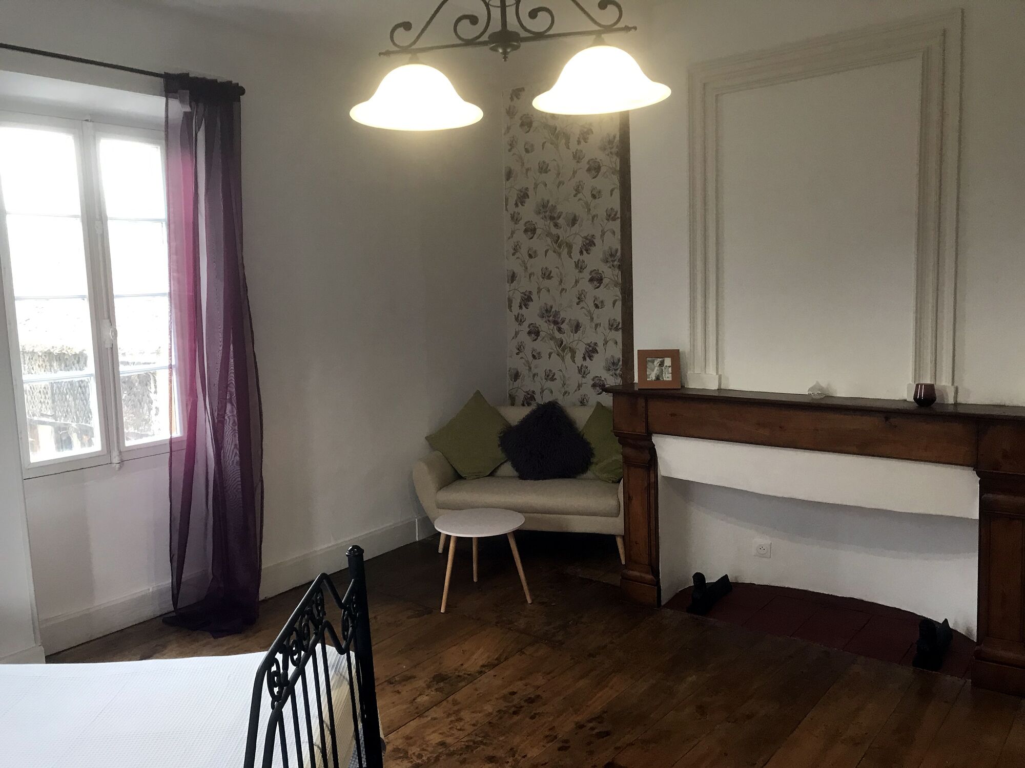 Een kamer met een houten vloer, een witte bank met kussens, een kleine ronde tafel, een zwarte metalen stoel, een open haard en een raam met gordijnen.