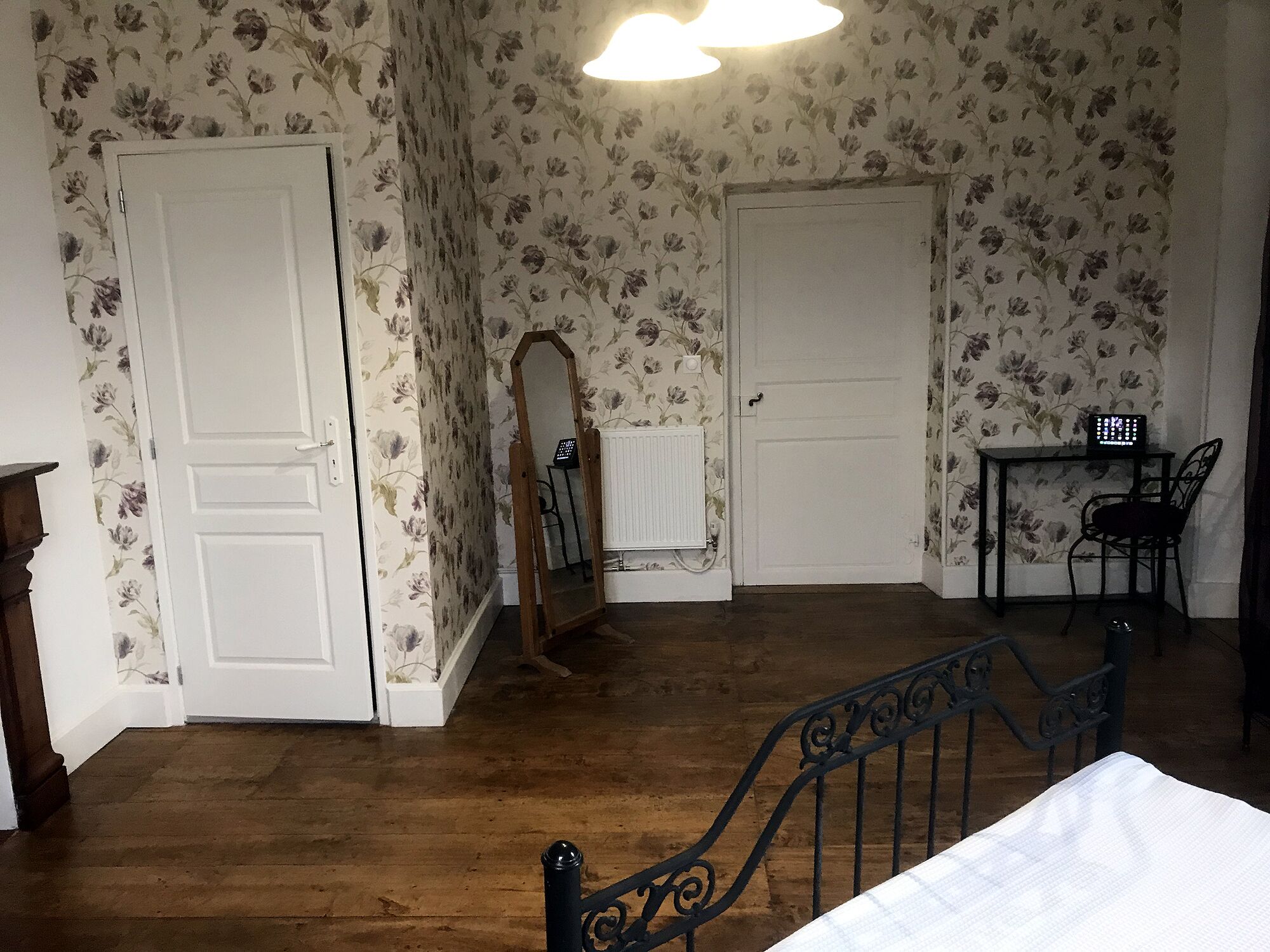 Una habitación con piso de madera, dos puertas blancas, un espejo, un radiador, una mesita con una silla y una cama con cubrecama blanco.