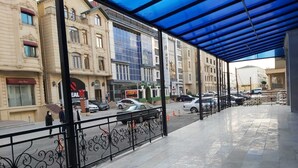 A hotel with a covered outdoor area, a tiled floor, and a view of the street with parked cars and buildings. - Baijan Hotel Baku (Baku)