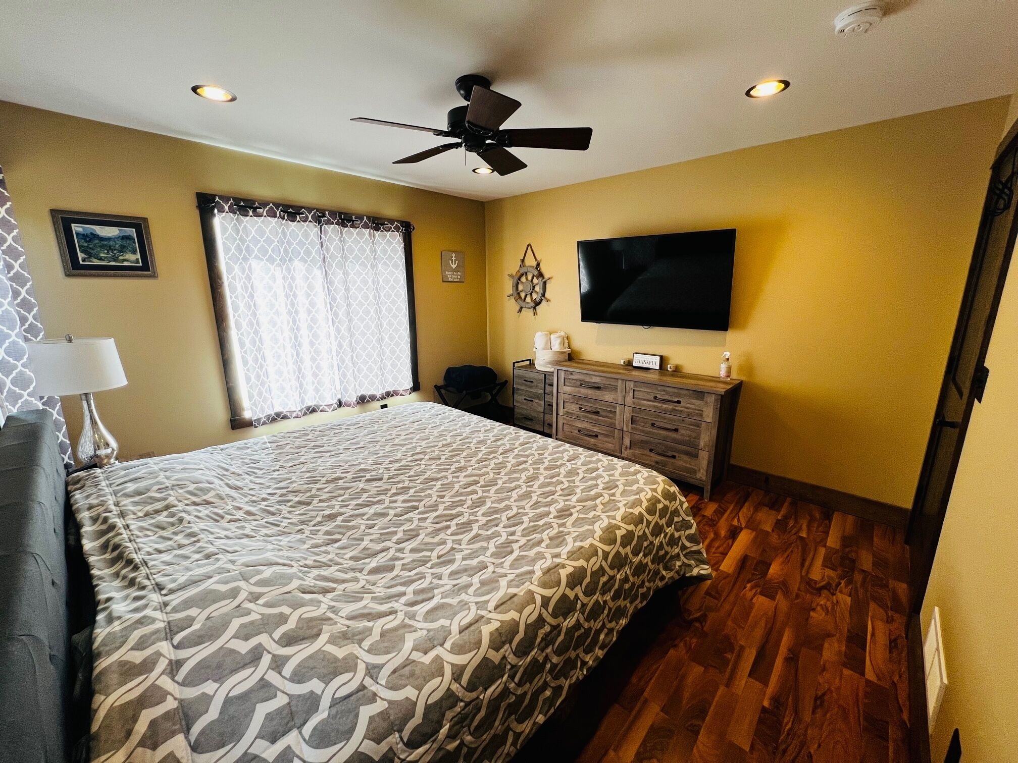 Quarto com cama, ventoinha no teto, televisão, cômoda e janela com cortinas.