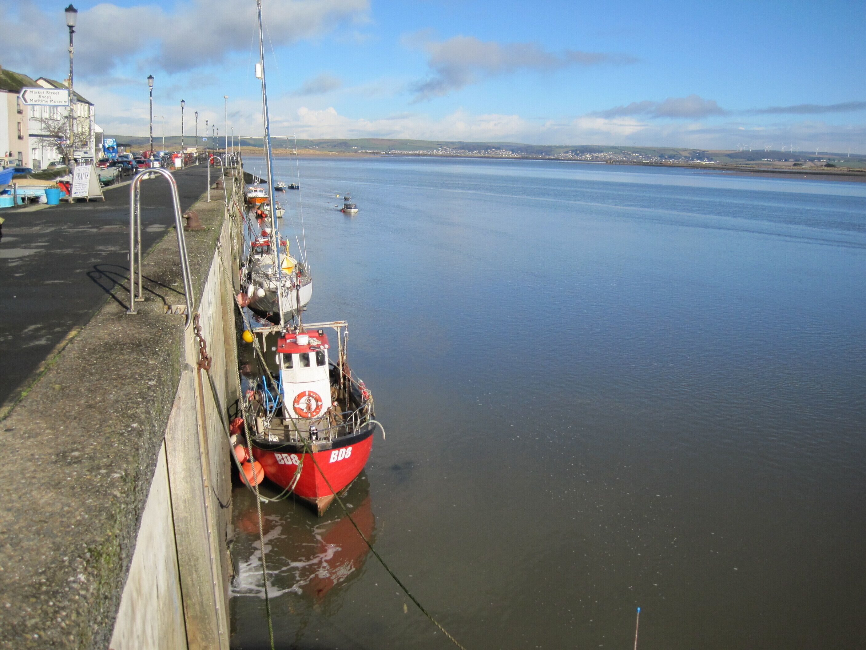 A harbor with boats docked along the quay, a clear sky, and distant wind turbines.