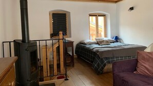 Tunnelmallinen huone, jossa on puukamiina, ruudullinen päiväpeitto, violetti sohva ja ikkunassa sälekaihtimet.