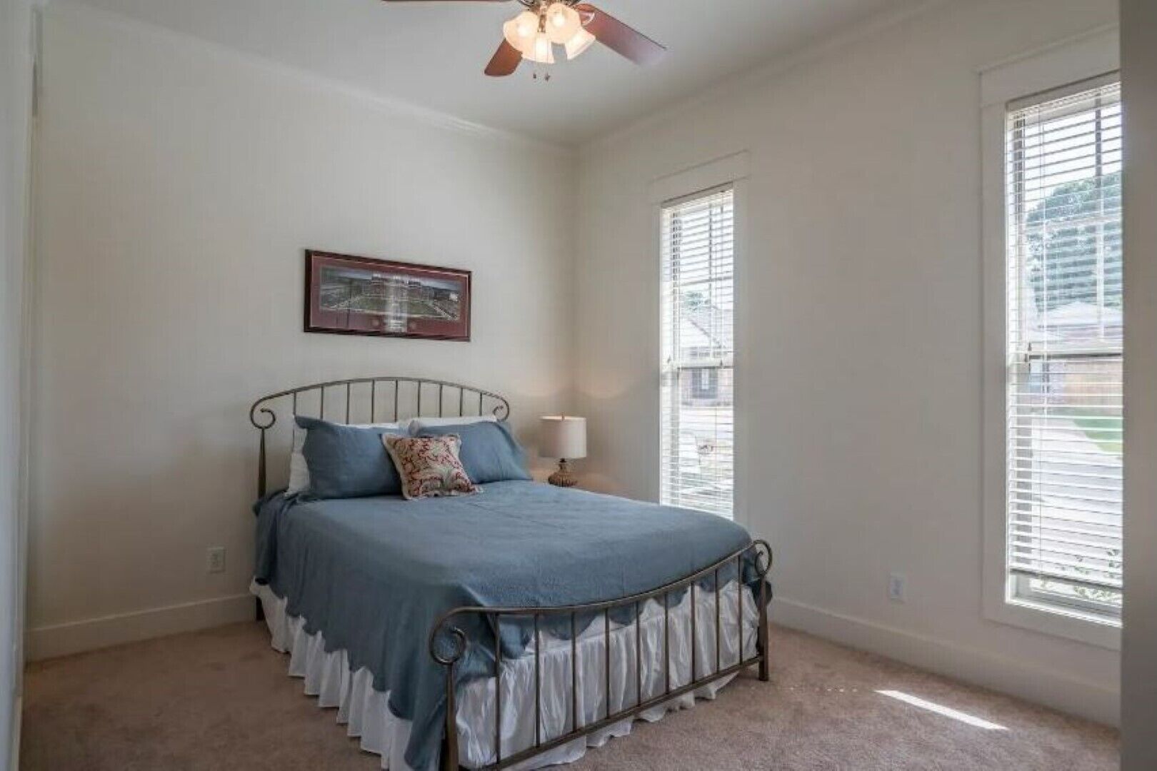 Une chambre à coucher avec un lit, un ventilateur de plafond, une fenêtre avec des stores et un tableau encadré au mur.