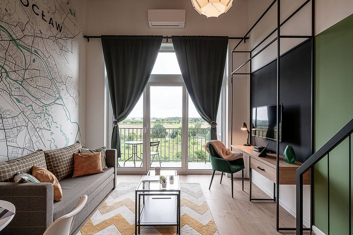 A modern living room with a sofa, a coffee table, a TV, and a balcony with a view of the outdoors.