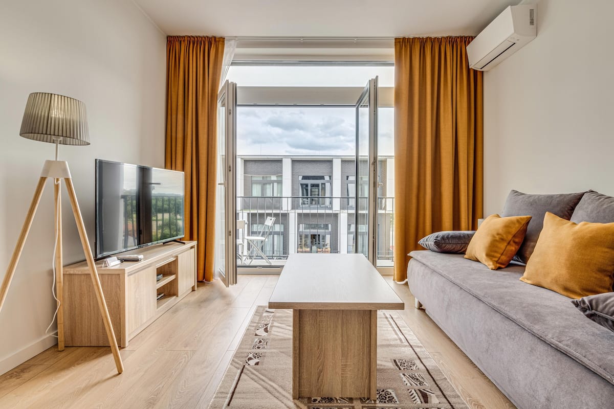 A modern living room with a flat-screen TV, a wooden coffee table, a grey sofa with cushions, and a large window with curtains.
