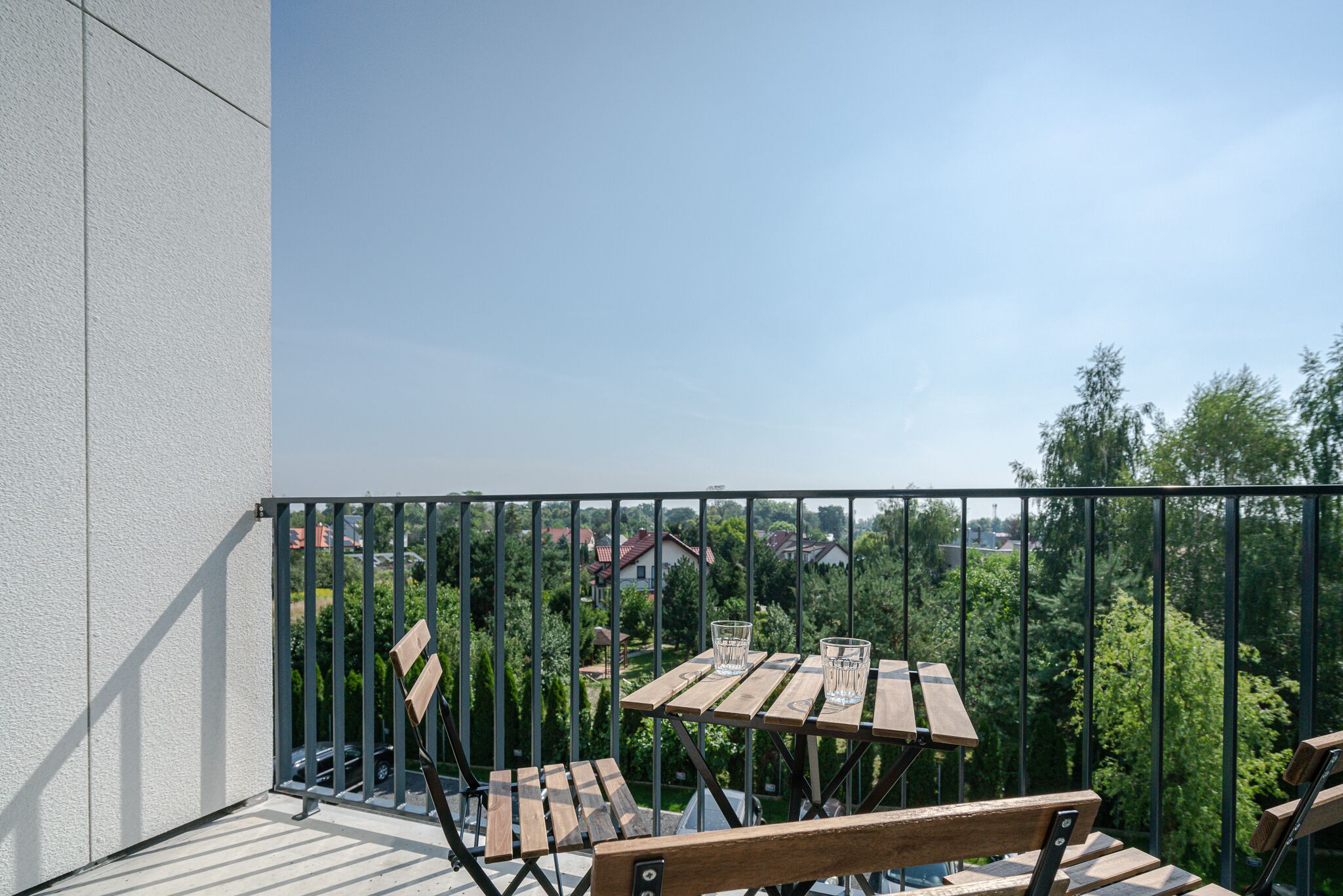 Een balkon met houten meubels en een tafel, met uitzicht op een woongebied met bomen en huizen.
