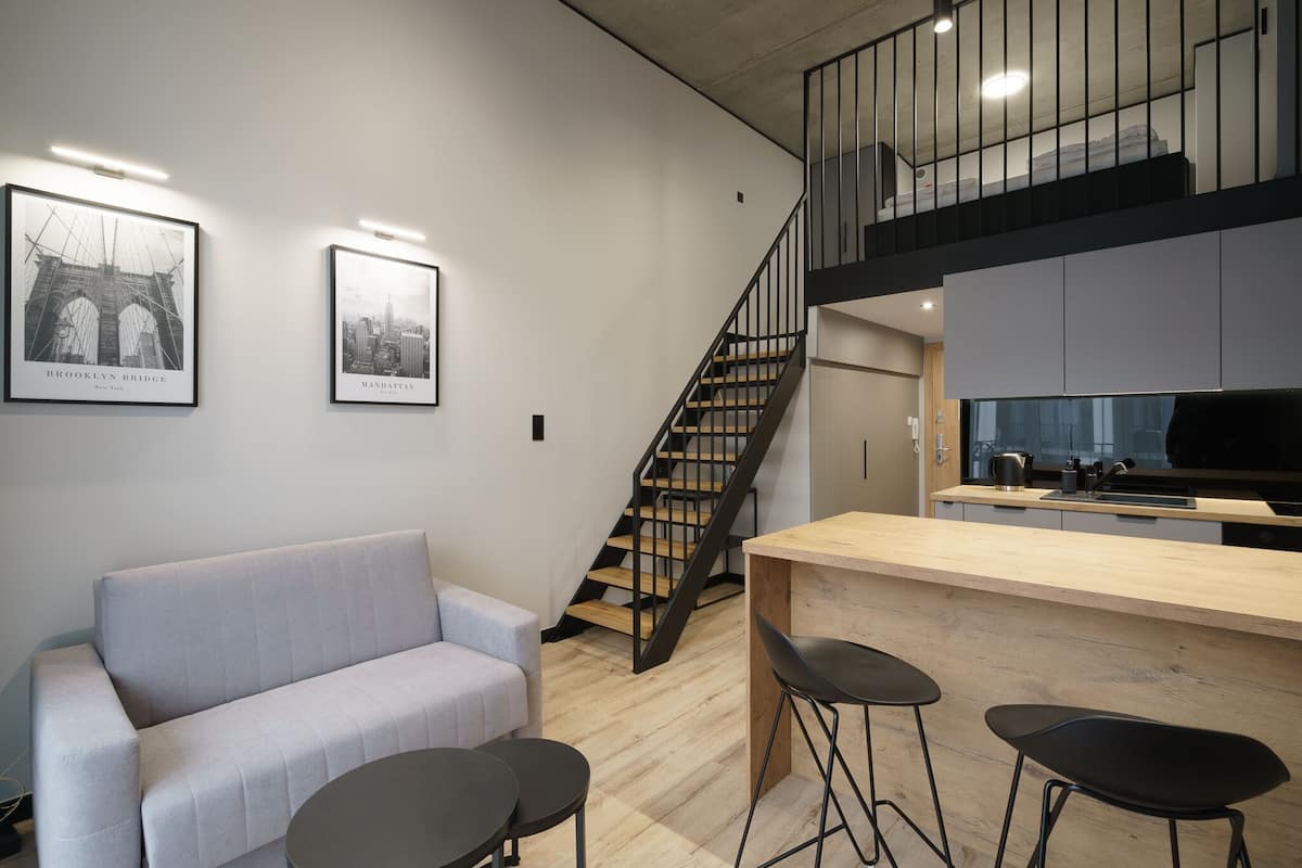 A modern apartment with a living area featuring a sofa and a dining area with a bar. A staircase leads to an upper level.