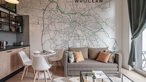 A modern living room with a large wall map of Wroclaw, a grey sofa with patterned cushions, white chairs, a glass coffee table, and a kitchen area with a black countertop and shelves. - RentPlanet - Apartamenty Zarembowicza (Wroclaw)