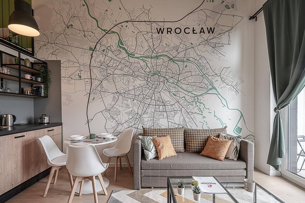 A modern living room with a large wall map of Wroclaw, a grey sofa with patterned cushions, white chairs, a glass coffee table, and a kitchen area with a black countertop and shelves.