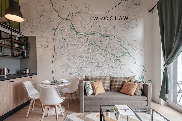 A modern living room with a large wall map of Wroclaw, a grey sofa with patterned cushions, white chairs, a glass coffee table, and a kitchen area with a black countertop and shelves. - RentPlanet - Apartamenty Zarembowicza (Wroclaw)
