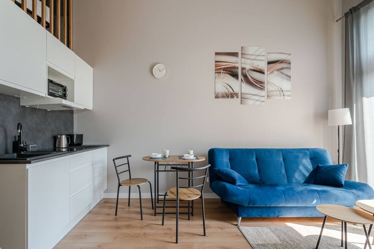 A modern living room with a blue sofa, a dining table with chairs, and a kitchen area with white cabinetry.