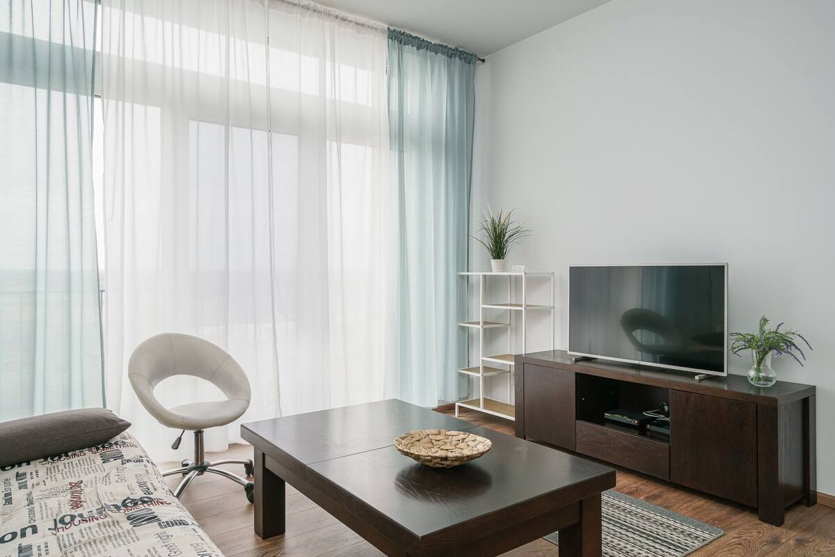 A modern living room with a dark wooden coffee table, a comfortable chair, a large flat-screen TV, and a bookshelf.