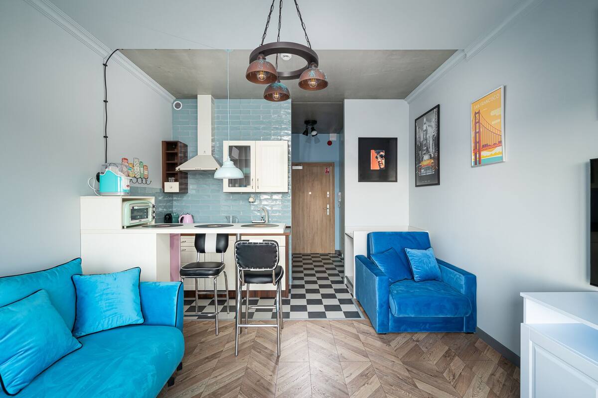 A compact living space with a kitchenette, a blue sofa, a blue armchair, and a herringbone-patterned floor.