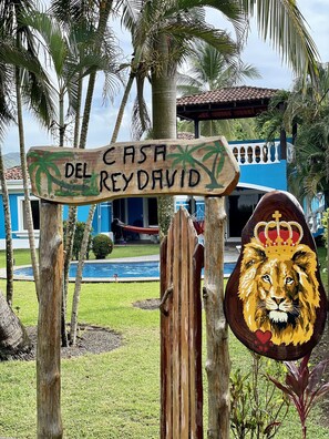 Une enseigne en bois sur laquelle est inscrit « Casa del Rey David », entourée de palmiers et bordée d’une piscine.