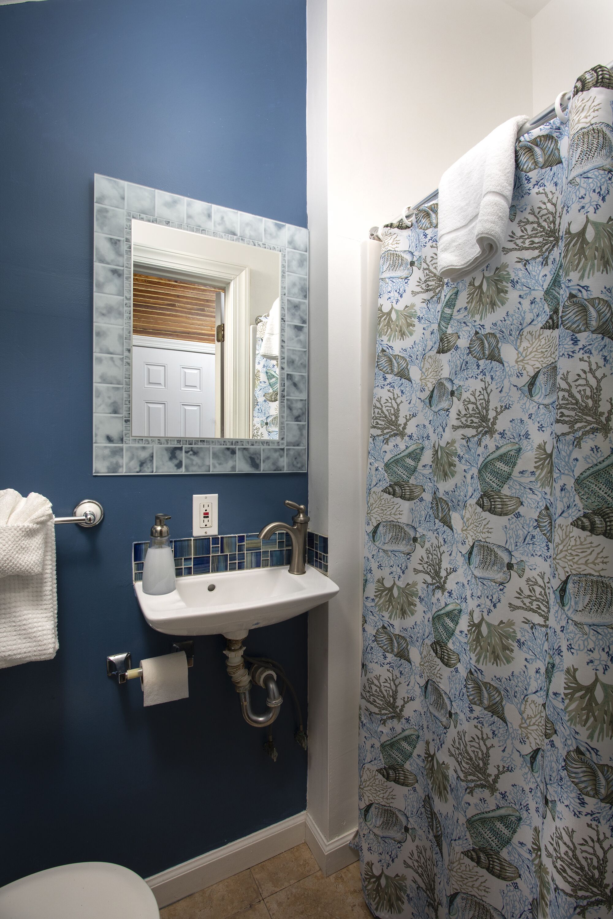 Une salle de bain avec un mur bleu, un lavabo blanc surmonté d’un miroir et un rideau de douche orné d’un motif représentant la vie marine.