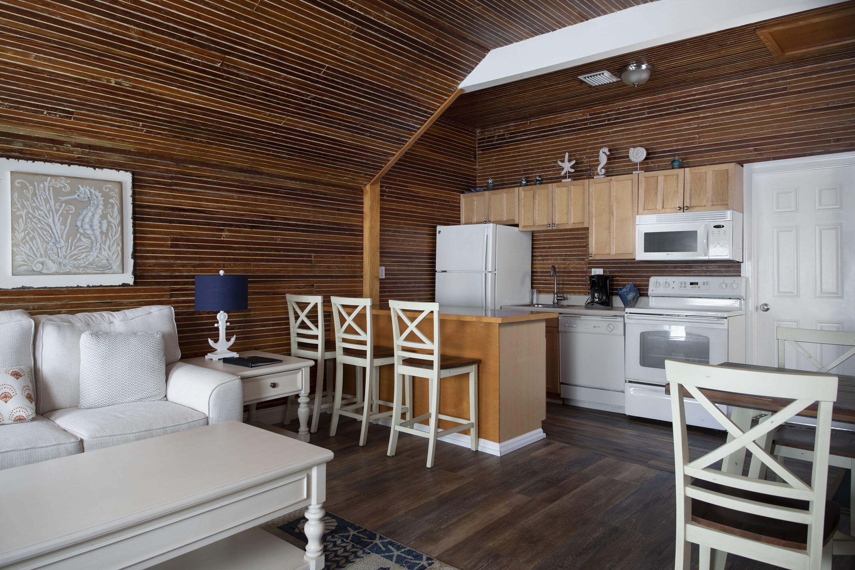 Una cocina compacta con paneles de madera, un refrigerador blanco, un microondas y una estufa. Hay un comedor con una mesa y sillas blancas.