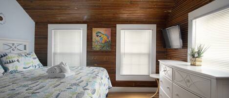 A bedroom with a wooden ceiling, a bed with a patterned quilt, two windows with blinds, a TV on the wall, and a dresser.
